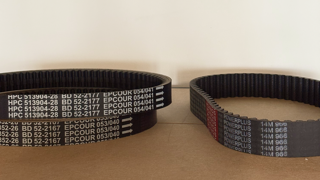 Three black and toothed belts on a wooden surface. The belt on the right reads "Movendis POWERPLUS."