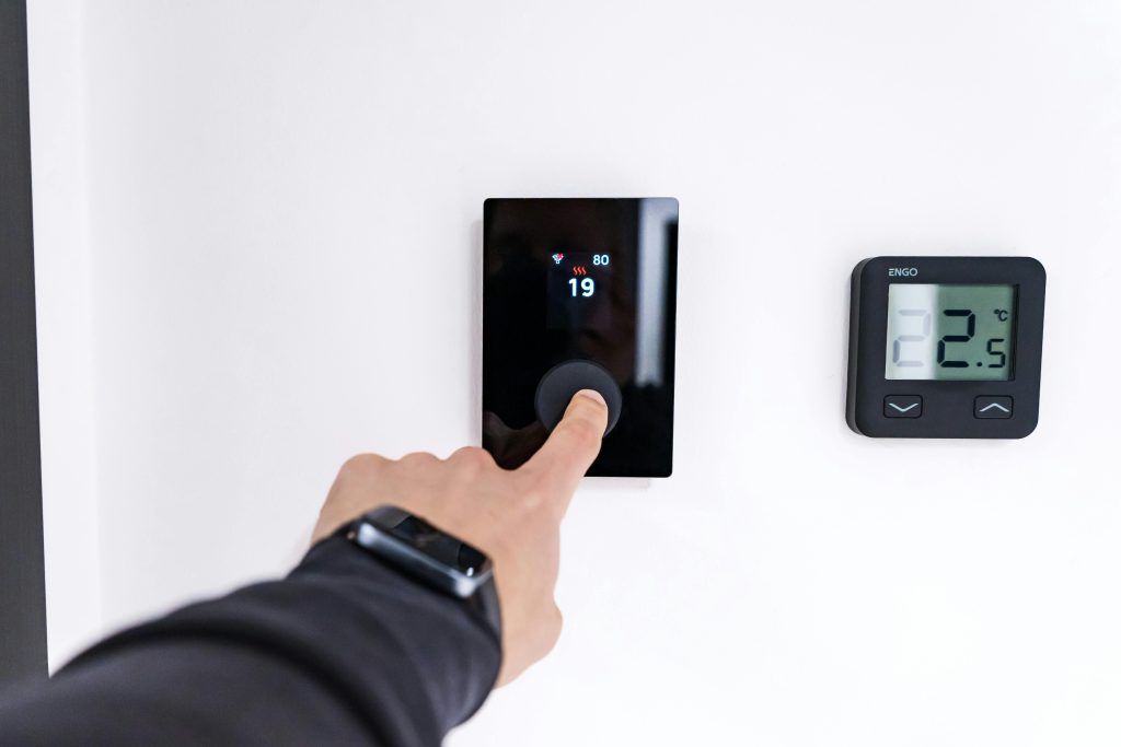 A person adjusts a digital thermostat on a wall, with their finger pressing a button. The screen shows "19" and another display reads "22.5°C."
